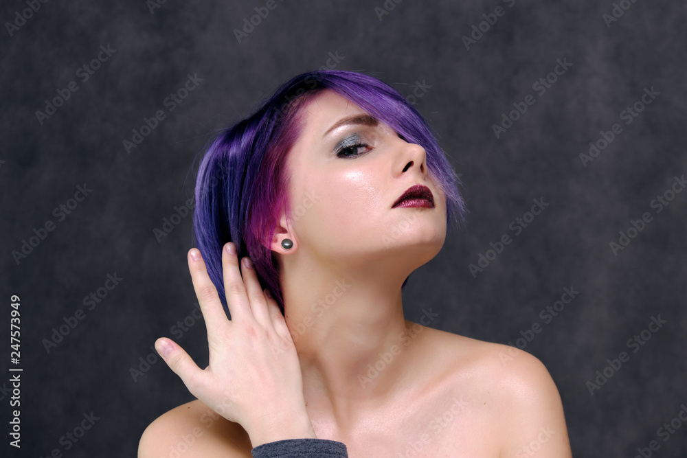 A beautiful, sexy girl with purple hair and a short haircut sits in the middle of the photo with a gray background