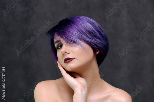 A beautiful, sexy girl with purple hair and a short haircut sits in the middle of the photo with a gray background