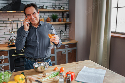 Happy adult man stand at table in kitchen and talk on phone. He hold glass of white wine and smile. Man cooking. Vegetalbes and cookbook on table. photo