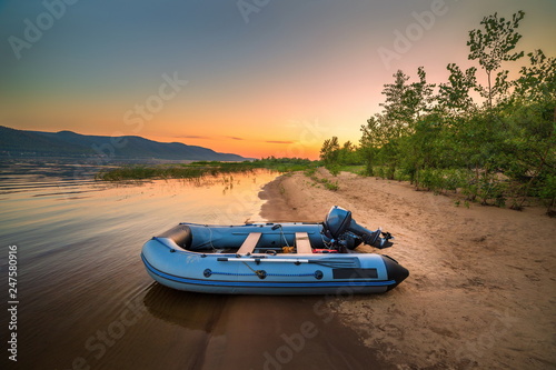 An inflatable boat with a motor on the shore of a sandy beach against the backdrop of a beautiful sunset.