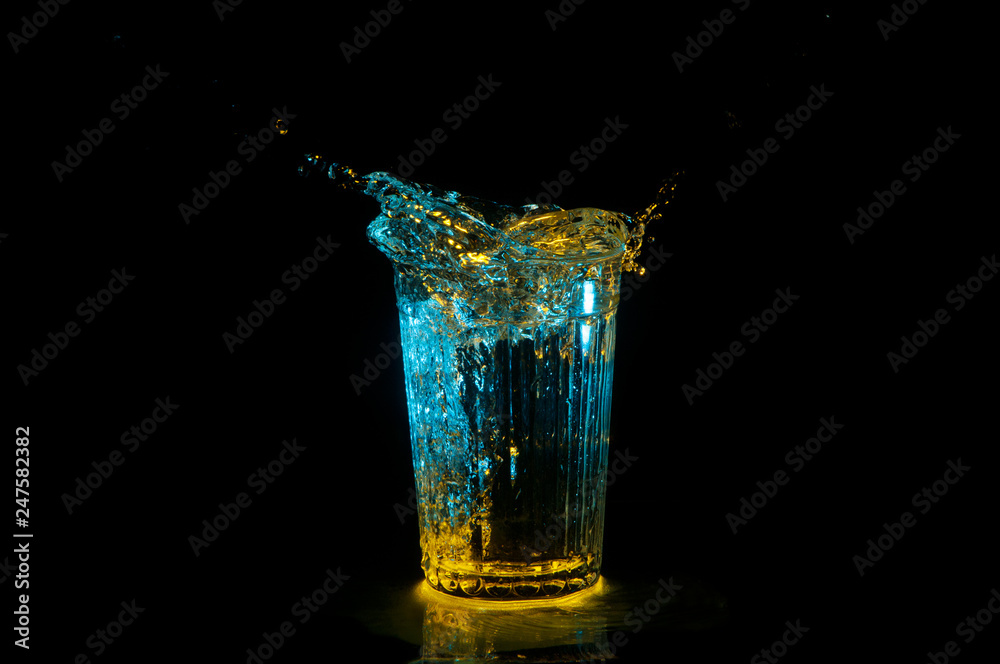 Ellipse of water splashing out of a ridged glass under yellow and blue lights isolated on a black background Ellipse of water splashing out of a ridged glass under yellow and blue lights isolated on a black background