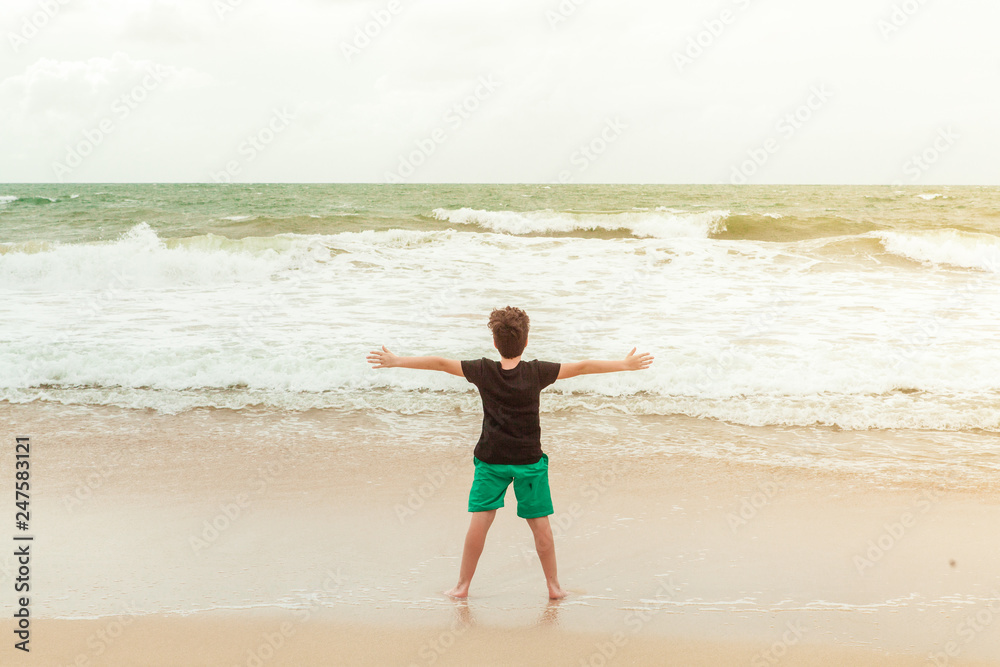 Boy in green shorts and black t-shirt standing by the ocean on the beach, looking at the waves and enjoying current moment Boy in green shorts and black t-shirt standing by the ocean on the beach, looking at the waves and enjoying current moment