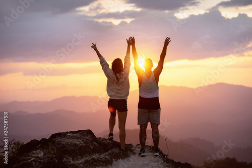 Happy couple with raised arms stands on mountain top against sunset and having fun in winner pose