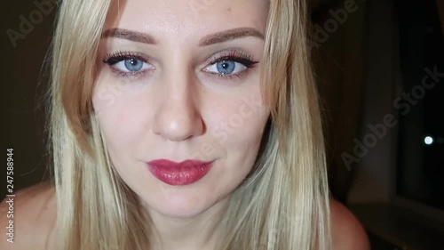 young beautiful blonde woman with bright blue big eyes and red lips flirts at the camera, raises one eyebrow and seduces with her eyes, close-up portrait on a dark background, slow motion