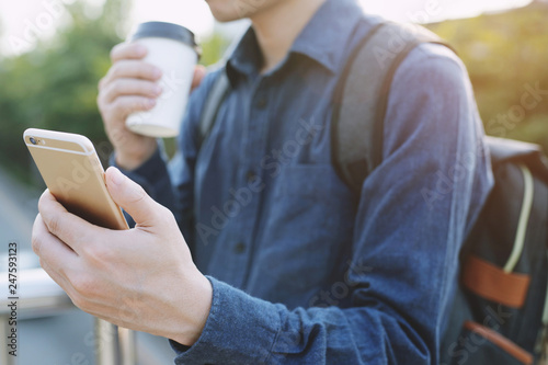 Travel blogger young man using route application using mobile smart phone reading to find the needed address in a city and holding a cup of take away coffee drink in morning sunlight. 