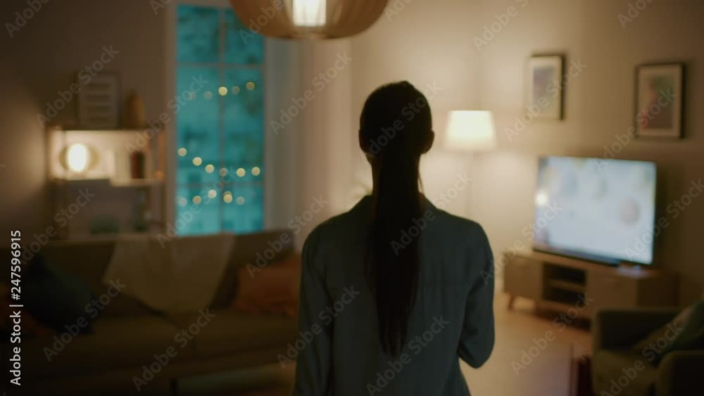 Young Beautiful Woman Gives a Voice Command to a Smart Home Application on Her Smartphone and Lights in the Room are Being Turned On. She Walks and Sits on a Couch. It's a Cozy Evening.