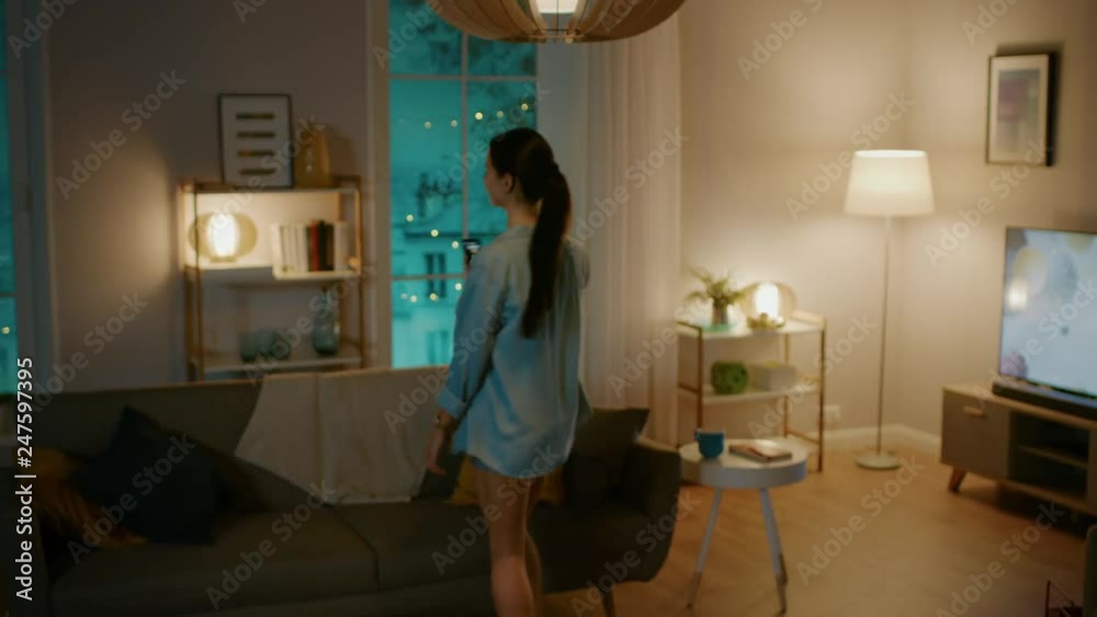 Young Beautiful Woman Gives a Voice Command to a Smart Home Application on Her Smartphone and Lights in the Room are Being Turned On. She Walks and Sits on a Couch. It's a Cozy Evening.