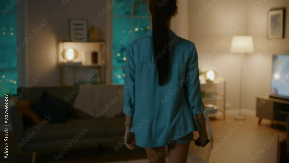 Young Beautiful Woman Gives a Voice Command to a Smart Home Application on Her Smartphone and Lights in the Room are Being Turned On. She Walks and Sits on a Couch. It's a Cozy Evening.
