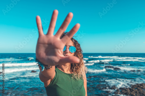 Trendy lady in sunglasses showing stop gesture to camera while standing near beautiful waving sea against cloudless blue sky on sunny day photo