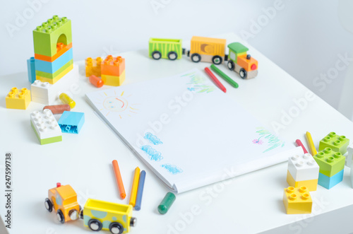 White table in the bright children's room with pencils, toy train, colorful plastic block and an album with children's drawings. Children's drawing table.