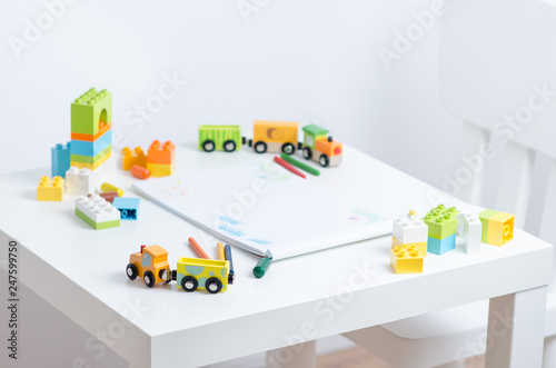 White table in the bright children's room with pencils, toy train, colorful plastic block and an album with children's drawings. Children's drawing table.