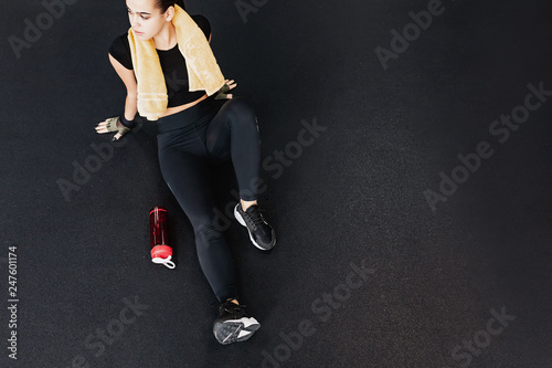 Strong but tired young strong-willed woman fitness trainer is tired after a grueling workout and rests on the black floor with a bottle of water. Concept of fitness and gaining muscle mass of women