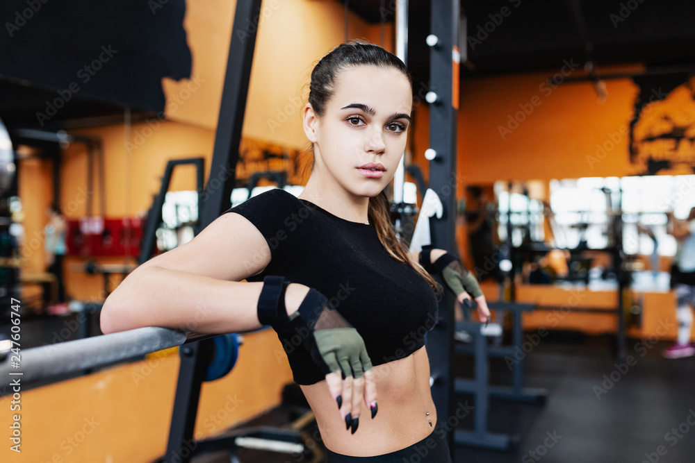 Beautiful young woman athlete fitness model in black sports top and leggings posing leaning on Smith's trainer. The concept of volitional discipline and endurance Beautiful young woman athlete fitness model in black sports top and leggings posing leaning on Smith's trainer. The concept of volitional discipline and endurance