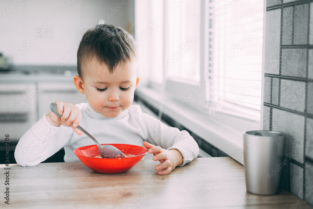 A child in the kitchen eating their own oatmeal with a red plate A child in the kitchen eating their own oatmeal with a red plate