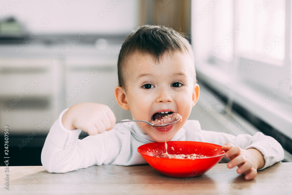 A child in the kitchen eating their own oatmeal with a red plate A child in the kitchen eating their own oatmeal with a red plate