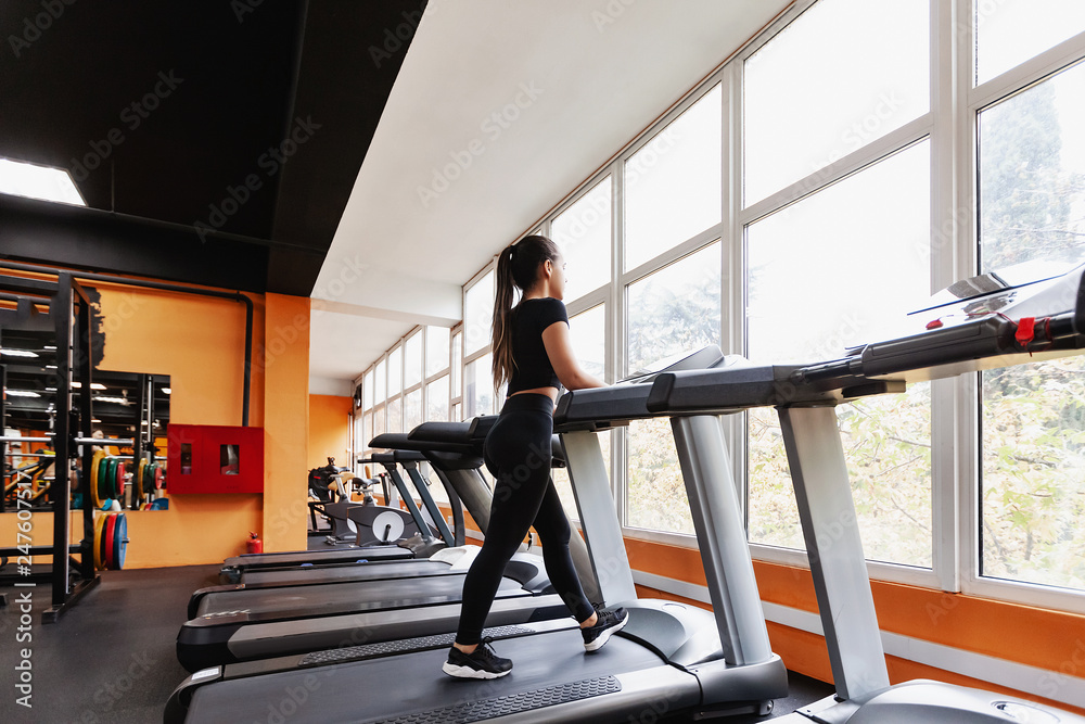 Obraz premium Slim young girl fitness model runs on a treadmill and looks out the window with a beautiful landscape. Concept of cardio for fat loss
