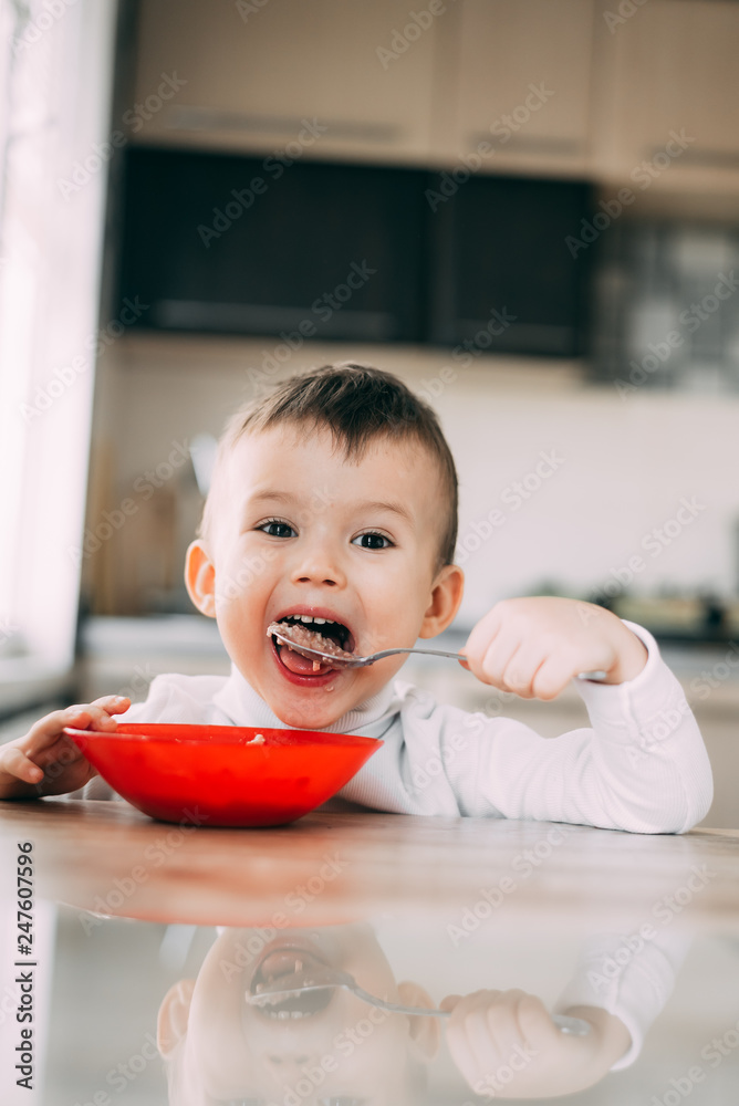 A child in the kitchen eating their own oatmeal with a red plate A child in the kitchen eating their own oatmeal with a red plate