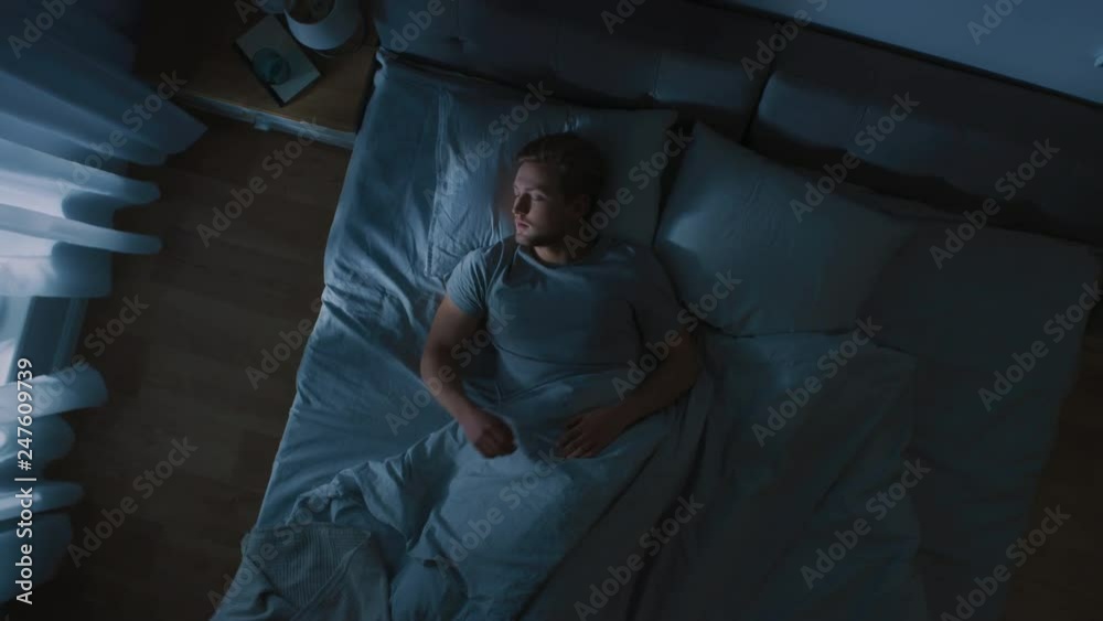 Top View of Handsome Young Man Sleeping Cozily on a Bed in His Bedroom at Night. Blue Nightly Colors with Cold Weak Lamppost Light Shining Through the Window.