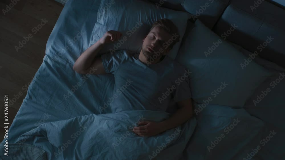 Top View of Handsome Young Man Sleeping Cozily on a Bed in His Bedroom at Night. Blue Nightly Colors with Cold Weak Lamppost Light Shining Through the Window.