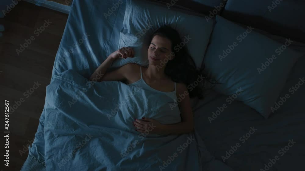 Top View of Beautiful Young Woman Sleeping Cozily on a Bed in His Bedroom at Night. Blue Nightly Colors with Cold Weak Lamppost Light Shining Through the Window.