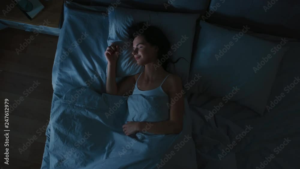 Top View of Beautiful Young Woman Sleeping Cozily on a Bed in His Bedroom at Night. Blue Nightly Colors with Cold Weak Lamppost Light Shining Through the Window.