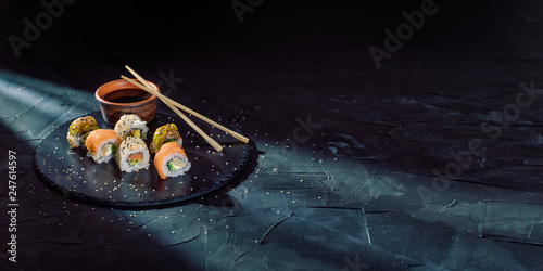 Quadro su tela A different assortment of sushi rolls on a black coal board and a dark background