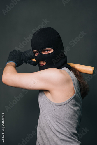 Bandit, the criminal in a black mask and in a gray T-shirt with a painless bat. Robber with a weapon. A man in a balaclava and a gray mask on a gray background