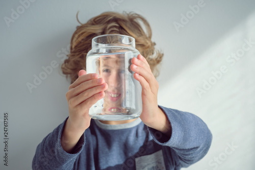 Cute little boy smiling and holding glass jar with clean water in front of face while standing near white wall photo