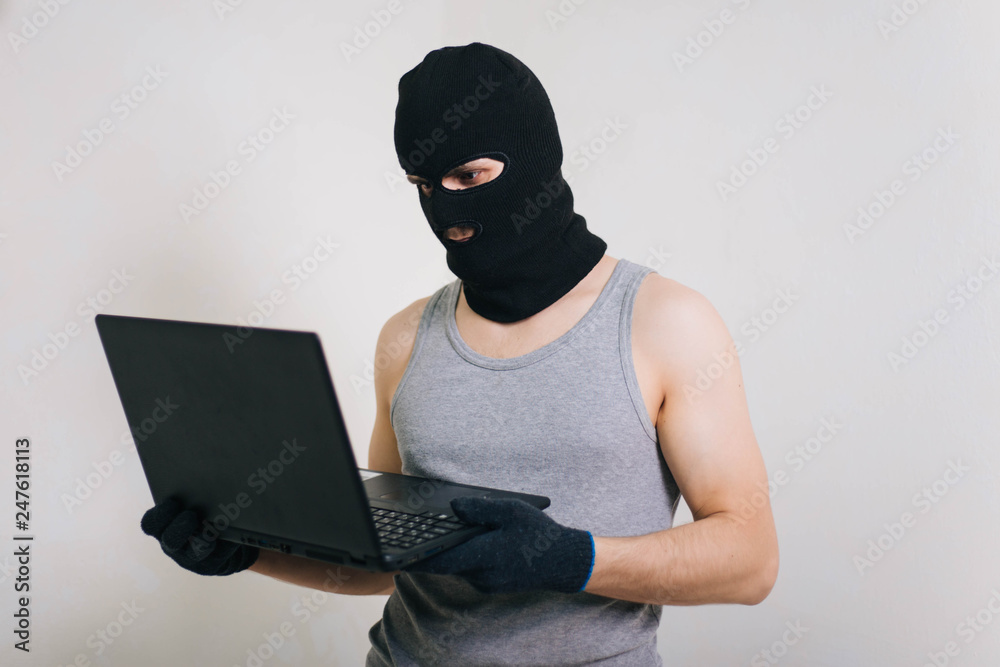 A thief in a black mask stole a laptop. A man in a balaclava and with a computer in hands on a white background. The hacker is hacking the computer. Steals information. A young man breaks the law. A thief in a black mask stole a laptop. A man in a balaclava and with a computer in hands on a white background. The hacker is hacking the computer. Steals information. A young man breaks the law.
