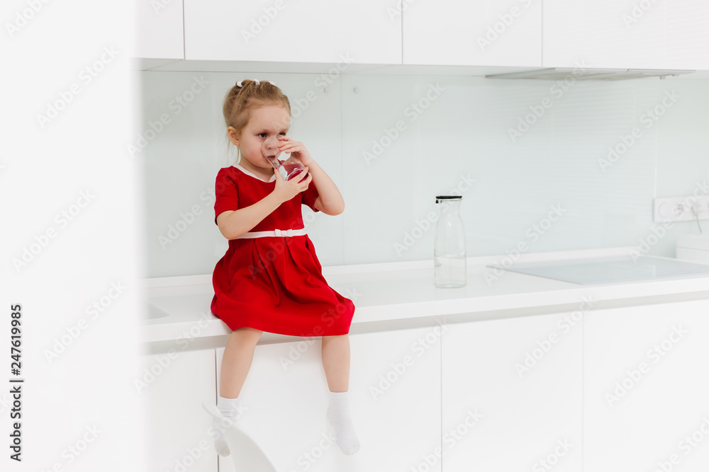 Cute clever little Caucasian girl in casual red dress holds a glass of water in her hands and drinks clear water, sits on a table in the white modern minimalistic kitchen. Cute clever little Caucasian girl in casual red dress holds a glass of water in her hands and drinks clear water, sits on a table in the white modern minimalistic kitchen.