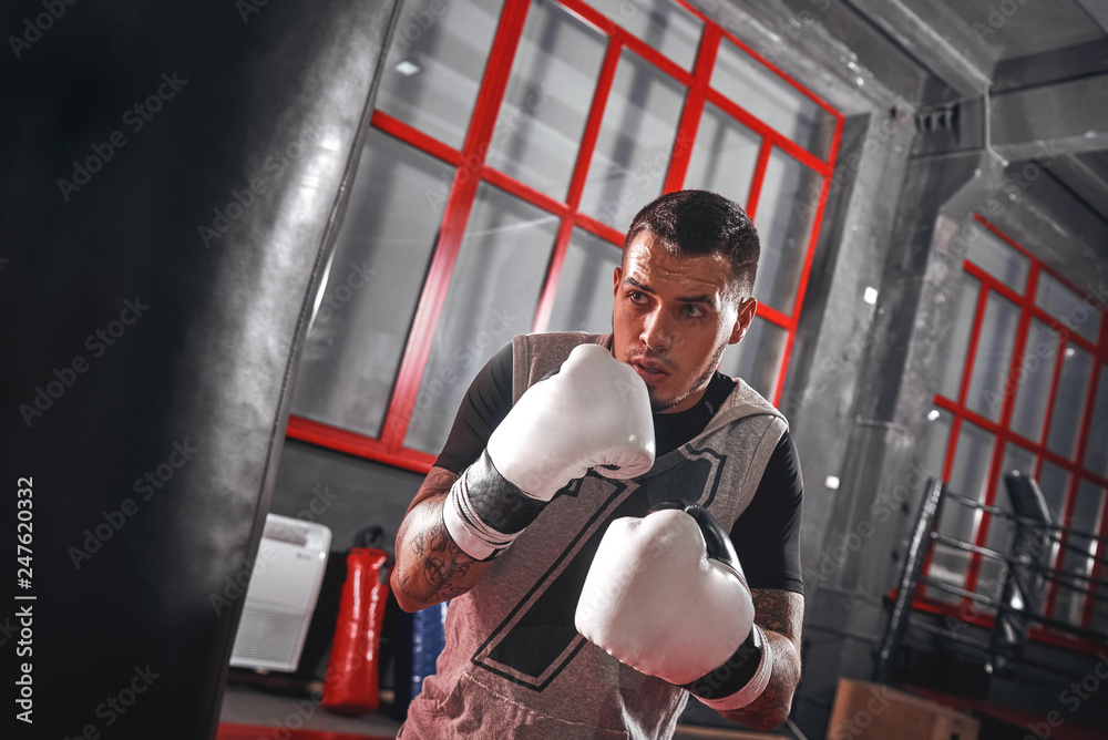 One second before. Focused tattooed athlete in sports clothing boxing on heavy punch bag while exercising in the boxing gym One second before. Focused tattooed athlete in sports clothing boxing on heavy punch bag while exercising in the boxing gym