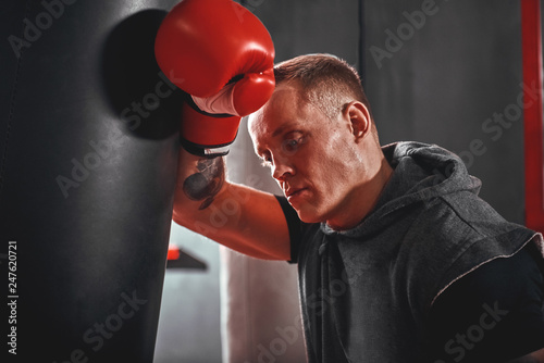 Tired after workout. Tired young athlete in sports clothing looking away after hard training on heavy punch bag in boxing gym