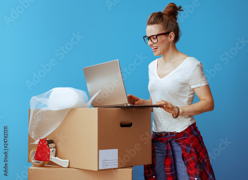 happy young woman near cardboard box using laptop on blue
