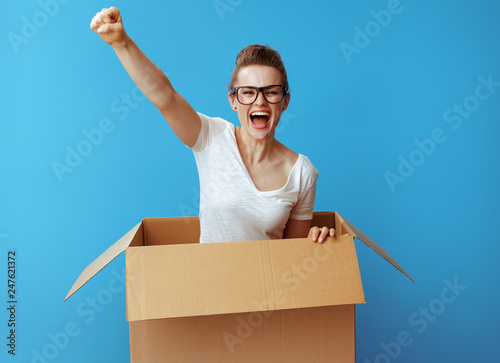 excited young woman pops out of cardboard box on blue
