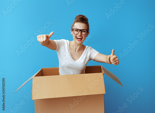 woman pops out of cardboard box and showing thumbs up on blue