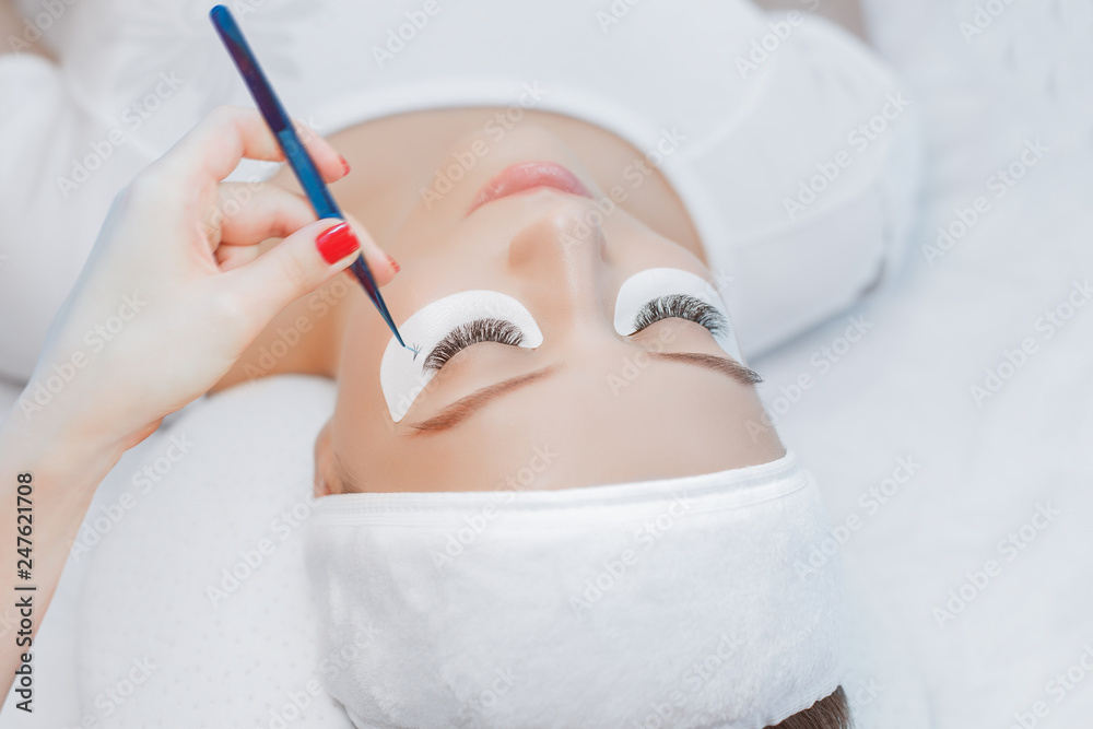 Naklejka premium Eyelash Extension Procedure. Close up view of beautiful Woman with Long Eyelashes. Stylist holding tweezers, tongs and making lengthening lashes for girl in a beauty salon. Beauty Concept.