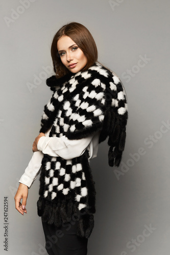 Fashionable and beautiful brown-haired model girl with perfect makeup in the black skirt and in a white blouse with trendy fur scarf posing in the studio at the grey background, isolated