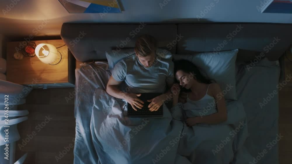 Top View Young Man in Bed Working on a Laptop Computer at Night while Girflfriend Sleeps Beside Him. Exceptionally Dedicated Project Manager Finishing Work in Bed at Night. Top View Young Man in Bed Working on a Laptop Computer at Night while Girflfriend Sleeps Beside Him. Exceptionally Dedicated Project Manager Finishing Work in Bed at Night.