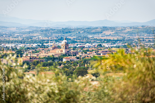 Many olive trees leaves framing town or village city of Bastia Umbra by Assisi with church in Umbria, Italy bokeh foreground during sunny summer day nobody