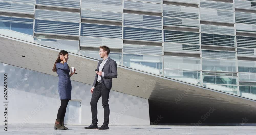 Smiling male and female colleagues talking while standing outside building. Businessman is holding mobile phone and businesswoman is holding disposable cup. They are wearing formals in city.