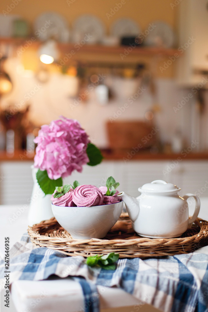 Pink marshmallows, white teapot and hydrangea flowers are serving on straw tray. Morning breakfast on kitchen table. Tea time, home atmosphere hygge. Cozy still life with sweets. Pink marshmallows, white teapot and hydrangea flowers are serving on straw tray. Morning breakfast on kitchen table. Tea time, home atmosphere hygge. Cozy still life with sweets.