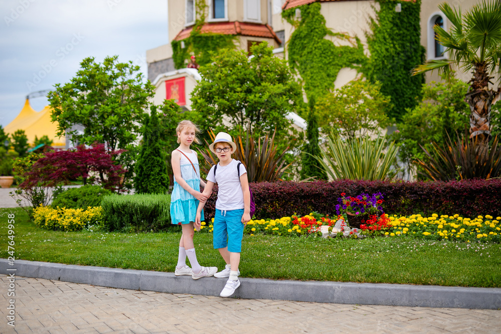 Obraz premium Blonde girl in white and blue dress and ginger boy in the hat and big glasses in the summer park with decorations