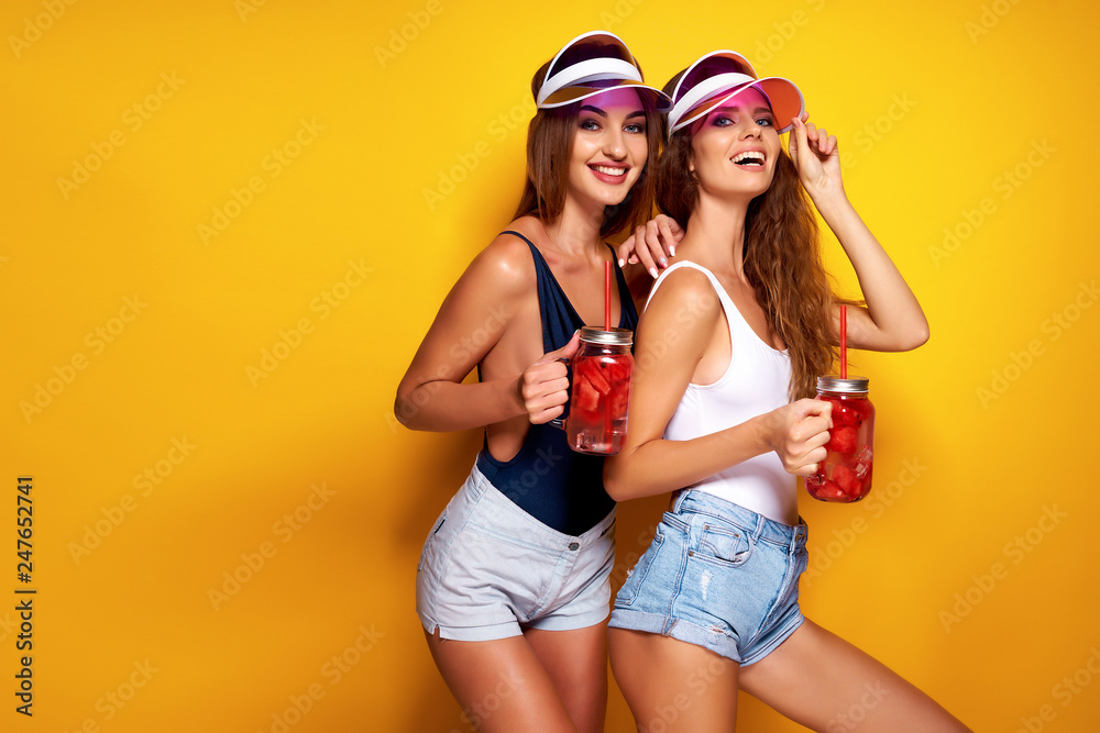 Two sensual young women in summer outfits, cap, swimsuits holding jars with fresh beverages while standing on bright yellow background - obrazy, fototapety, plakaty 