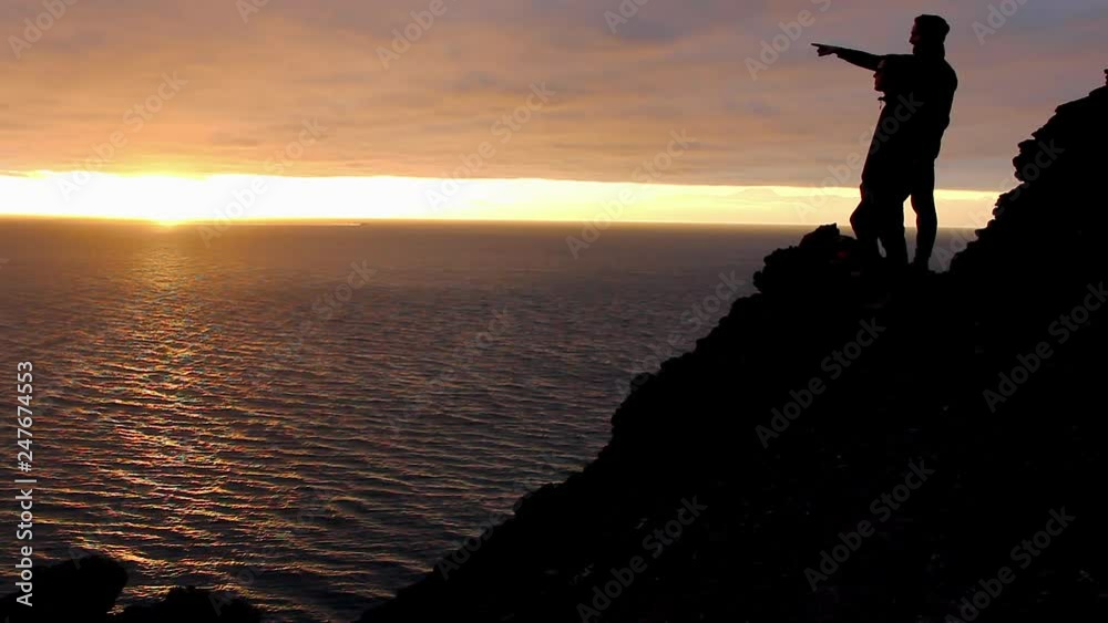 Young man pointing at horizon explaining splendid landscape by the sea at sunset in Gran Canaria, Spain. Silhouettes of friends standing on cliff edge with Teide mountain on background at twilight