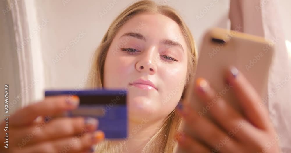A blonde girl sits with a telephone in her room amid pink curtains and garlands and scrolls the news line on the phone in search of the right product. A blonde girl sits with a telephone in her room amid pink curtains and garlands and scrolls the news line on the phone in search of the right product.