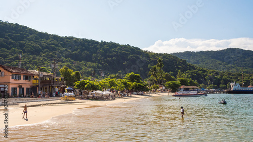 Rio de Janeiro / Brazil: February 20 2016:Village by the sea with a tourist beach, people on the sand and the warm crystal water with tropical jungle behind with a pier and boats on a summer sunny day