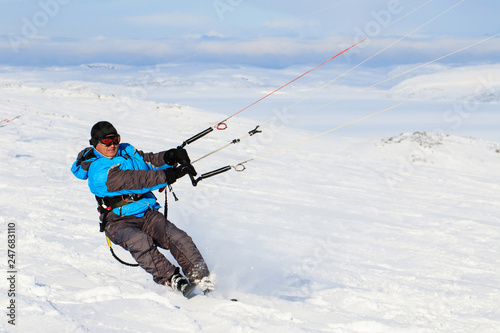 Man kite boarding rider sportsman with kite in sky rides in snow on kite board. Recreational activity, extreme active sports, snow kiting ski, outdoor, winter snowy season, skiing. Blue sky background