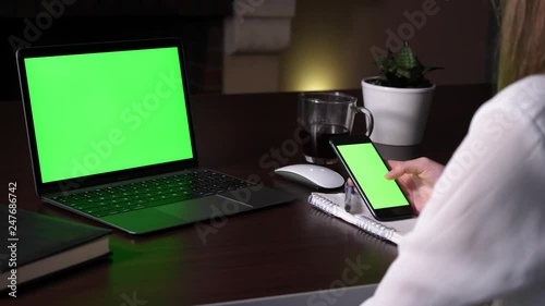 Woman in white shirt scrolls an application (green screen) on her smartphone in front of open laptop with green screen