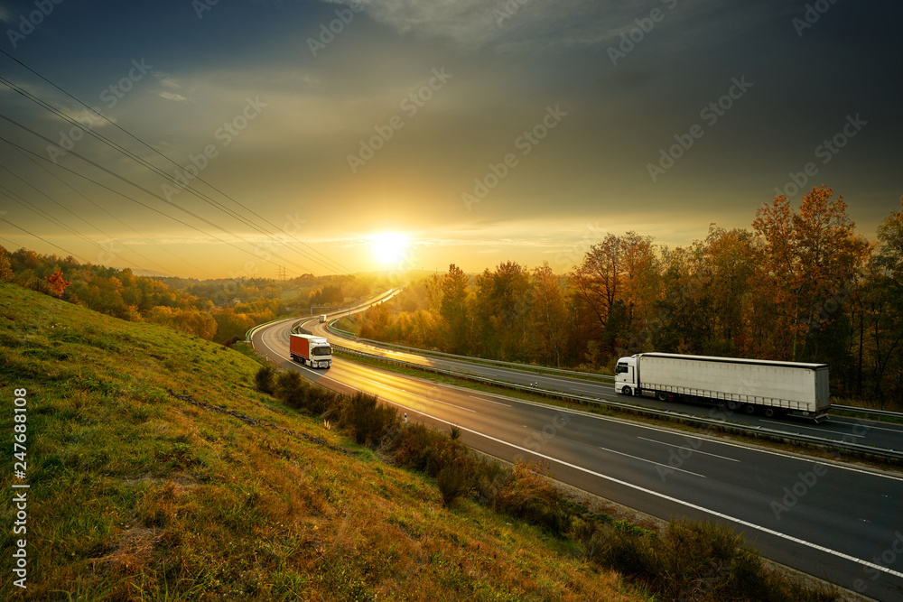 Three trucks driving on the asphalt highway in autumn landscape at sunset