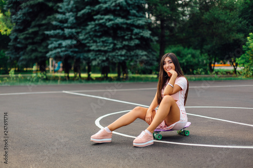 Portrait of a smiling charming brunette female sitting on her skateboard on a basketball court.